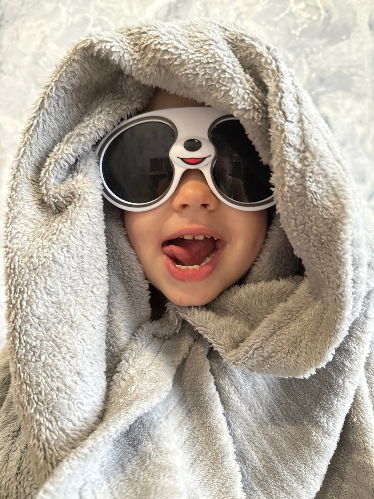 Child in novelty sunglasses lies wrapped in blanket indoors