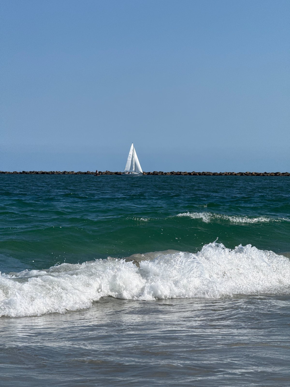 Sailboat cruising past Wrightsville Beach breakwater on calm day