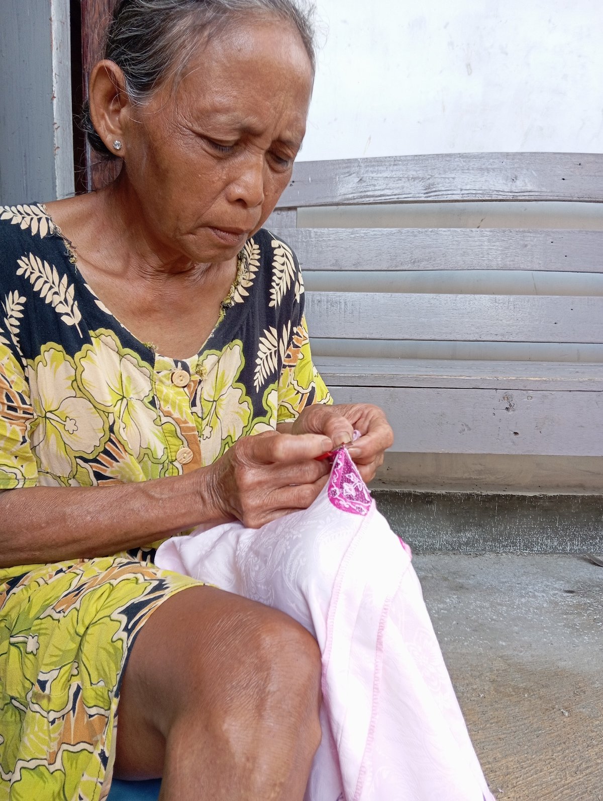 Elderly woman mends pink cloth outdoors in Baron, Indonesia