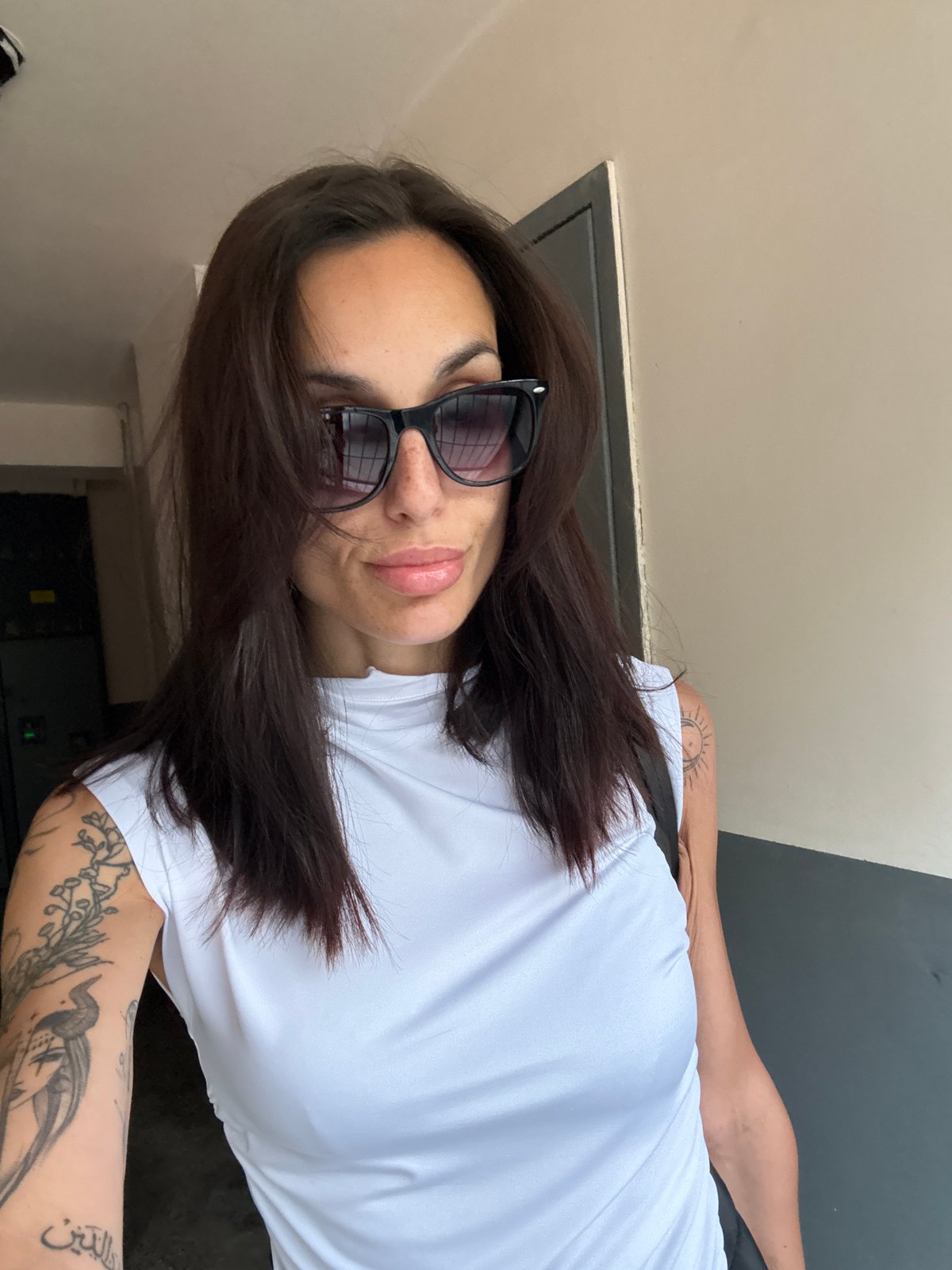 Tattooed woman in sunglasses observed against gray wall, Pleven