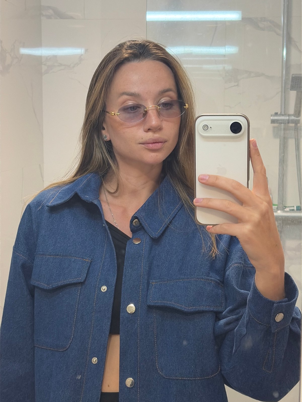 Woman takes bathroom selfie wearing denim jacket and glasses