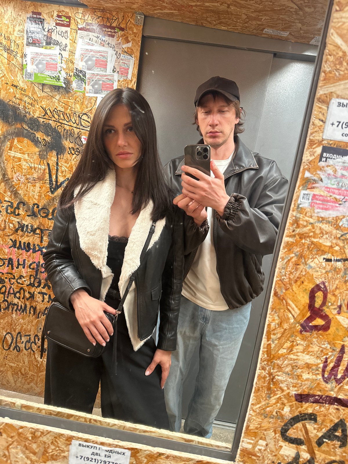 Couple takes elevator mirror selfie, woman in leather jacket