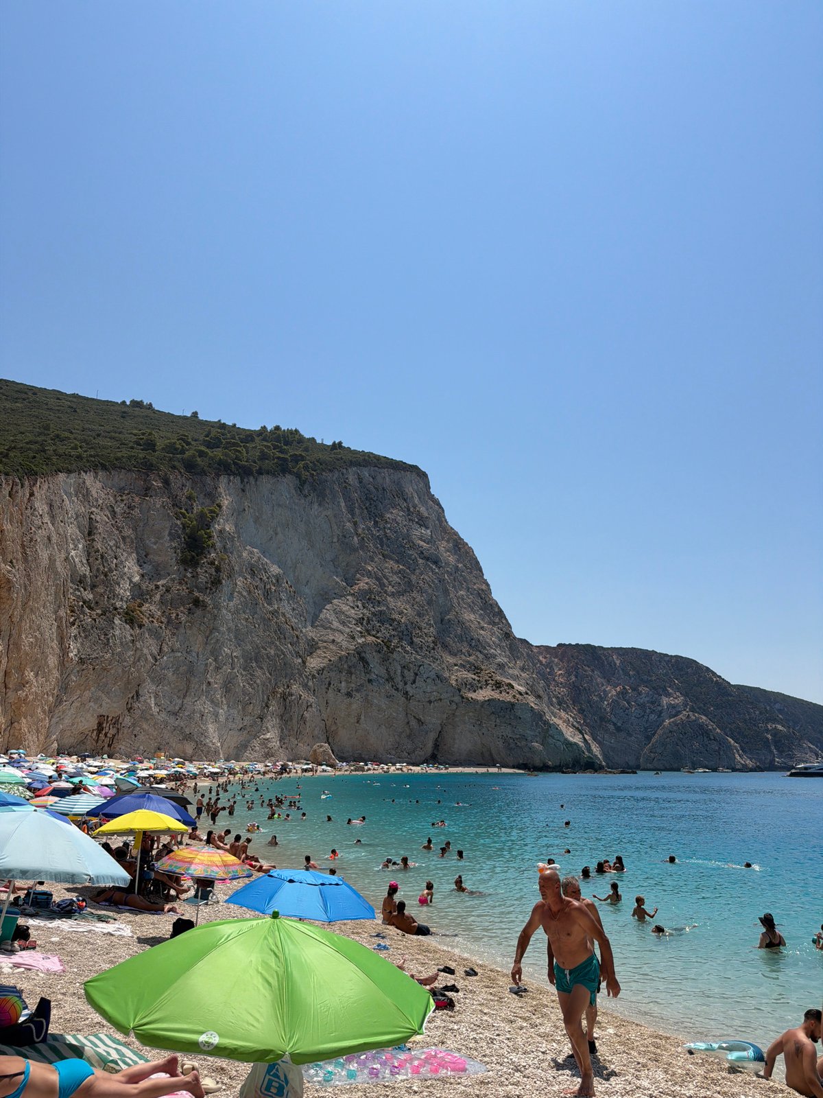Crowded pebble beach beneath a dramatic cliff, Apollonioi, Greece