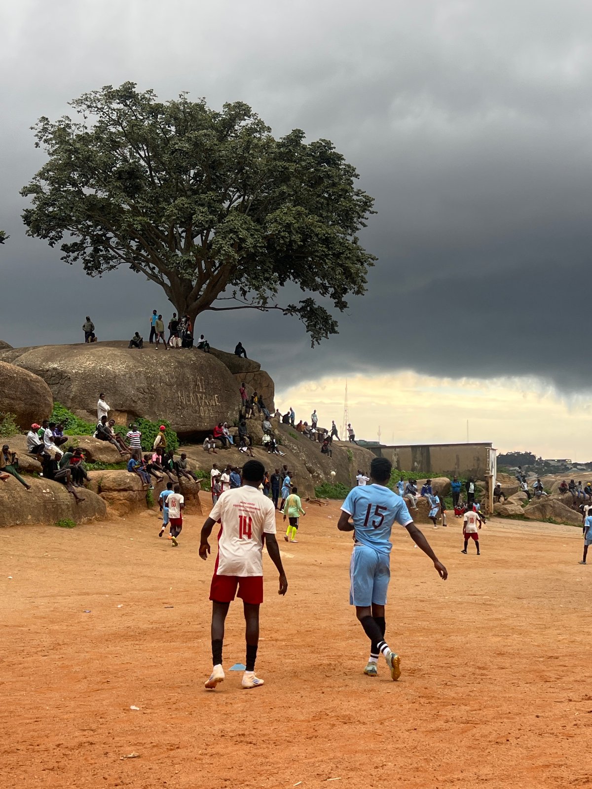 Football match on a dusty Jos pitch under gathering storm clouds