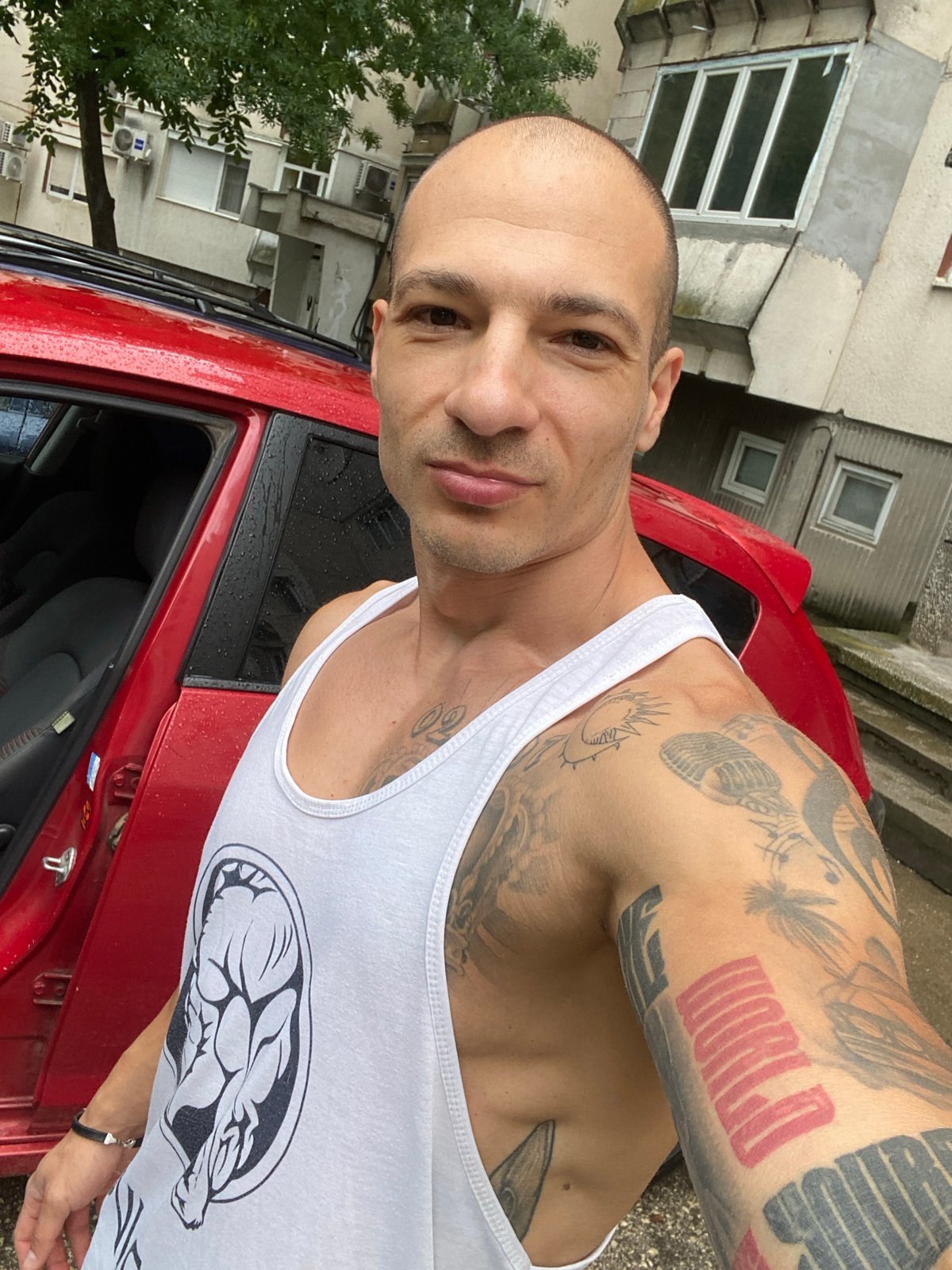 Tattooed arms, graphic tank, red car door open in Pleven