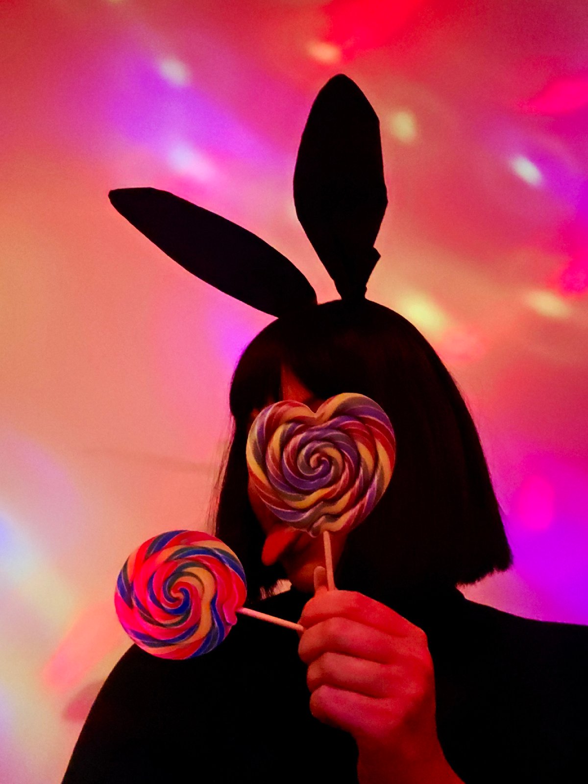 Person in bunny ears holds lollipops under colorful lights