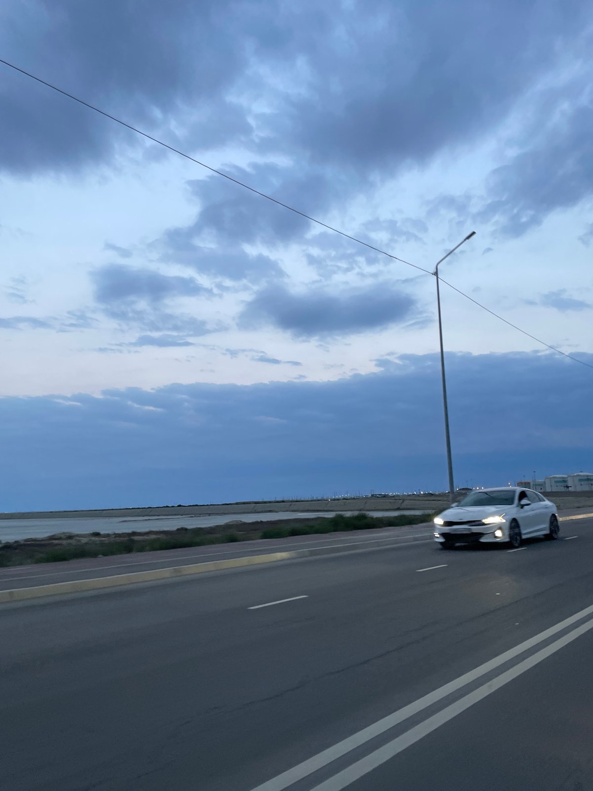 White sedan on a stormy Kazakh road, headlights cutting through overcast skies.