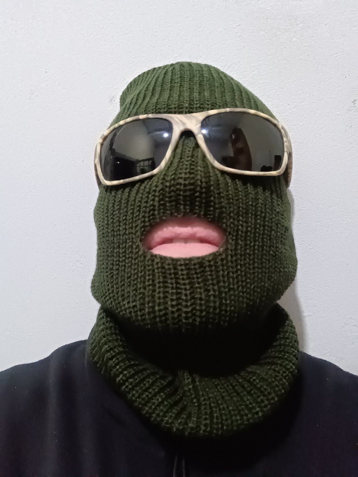 Masked individual in balaclava observed against grey wall