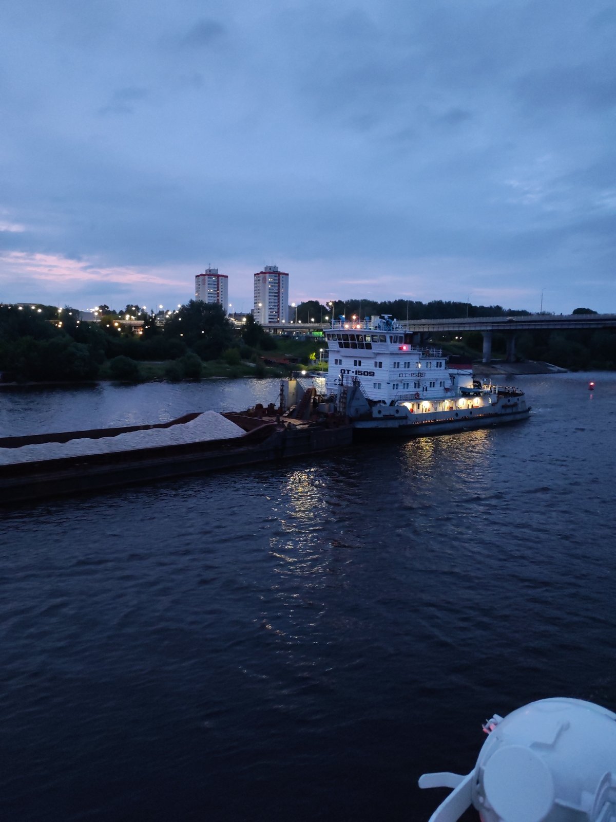 Evening river transit past lit bridge, barges, and industrial waterfront