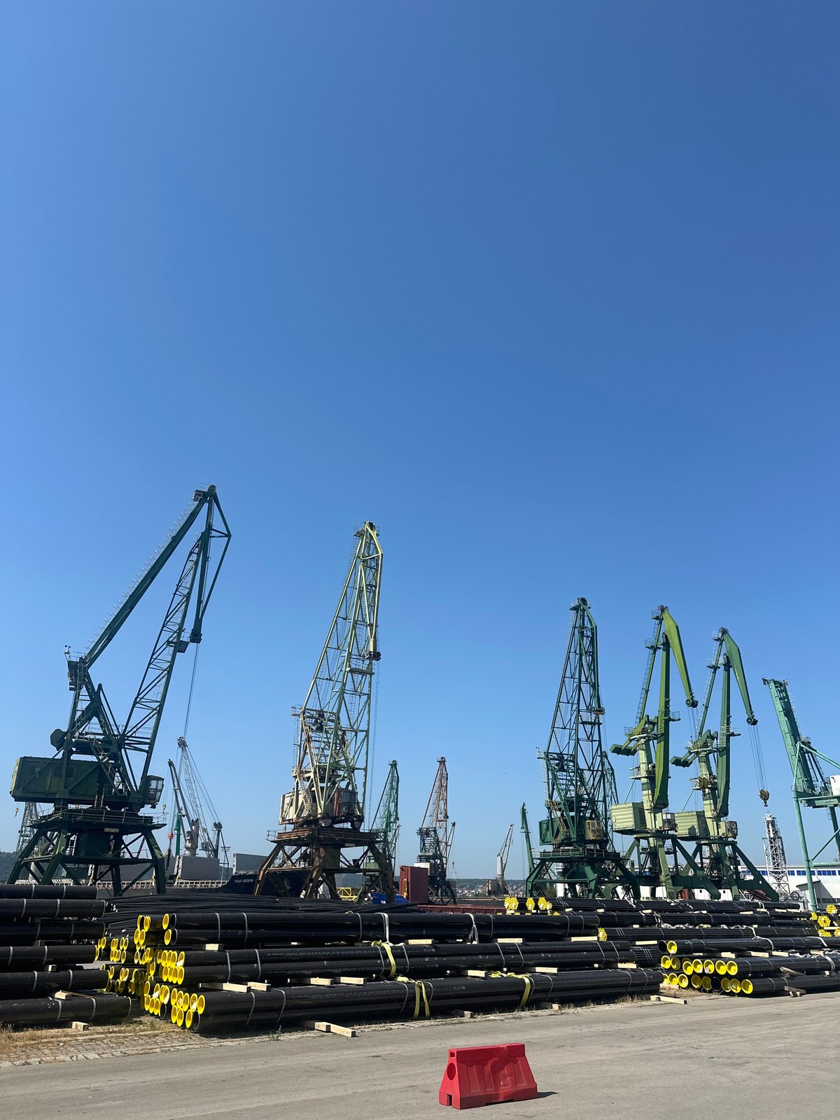 Industrial port yard with cranes, stacked pipes, and city backdrop
