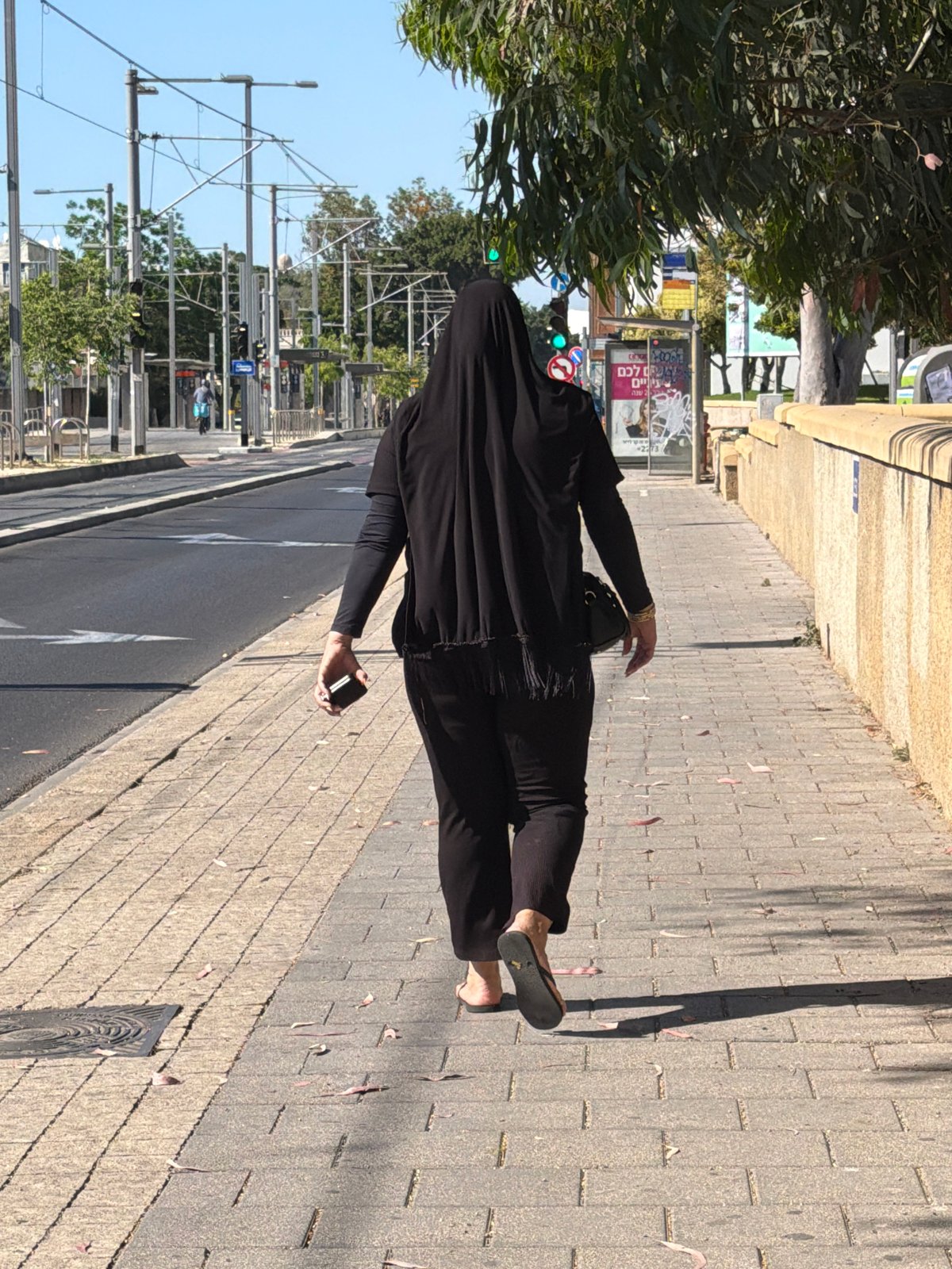 Person in black walks sidewalk with phone in Bat Yam