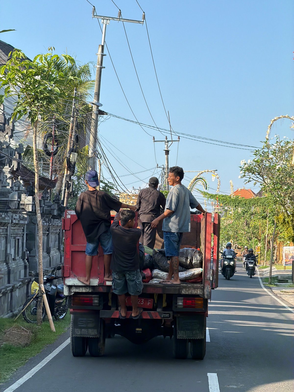 Red flatbed truck carries passengers through sunny Mengwi streets