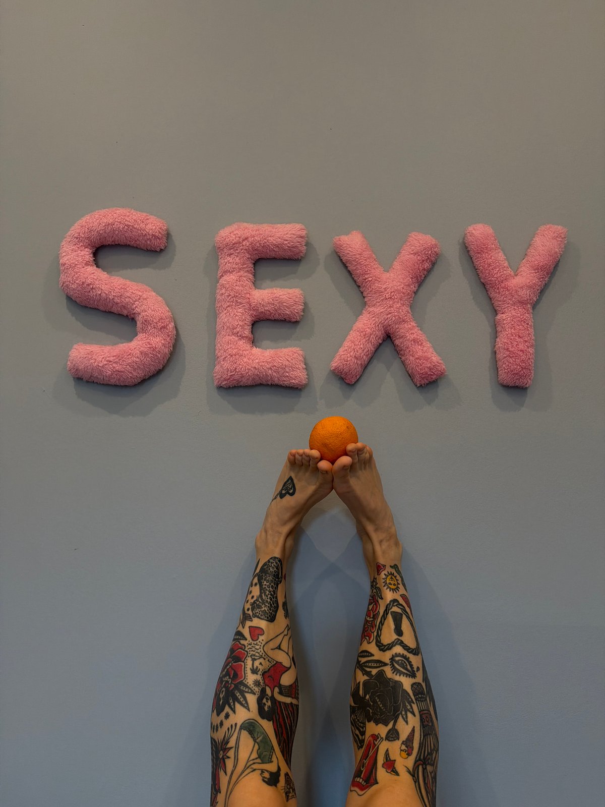 Tattooed feet, gray wall, pink fuzzy letters spelling SEXY