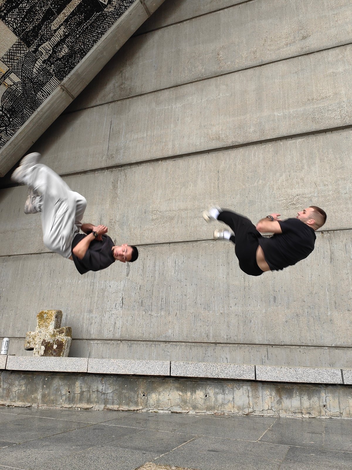 Two men perform synchronized aerial acrobatics near concrete structure