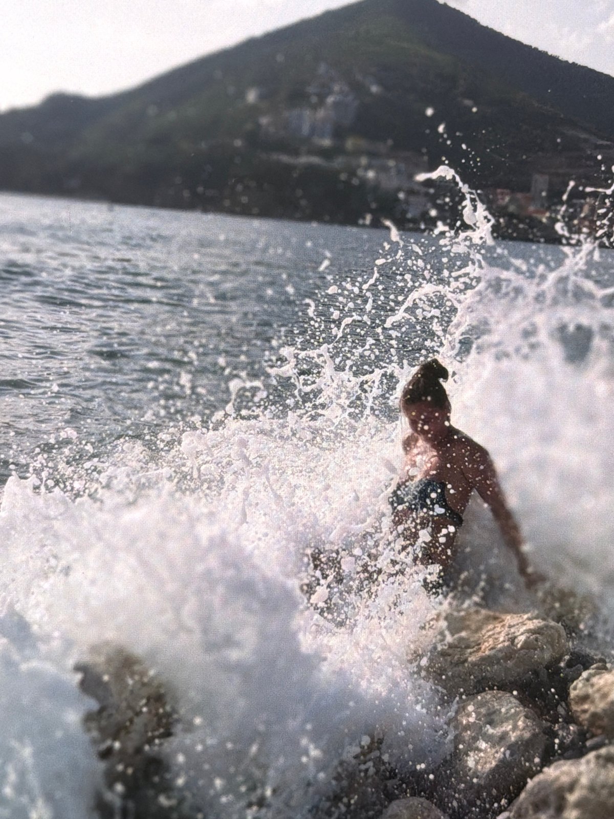 Waves hit a rocky shore near a quiet harbor