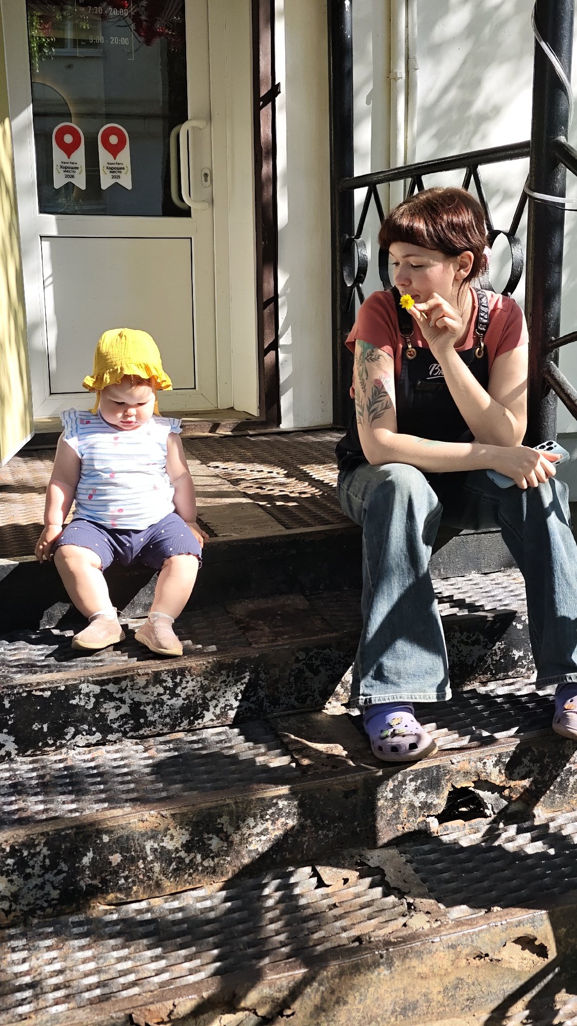 Mother, baby rest outside Chaikovsky business; person holds dandelion nearby