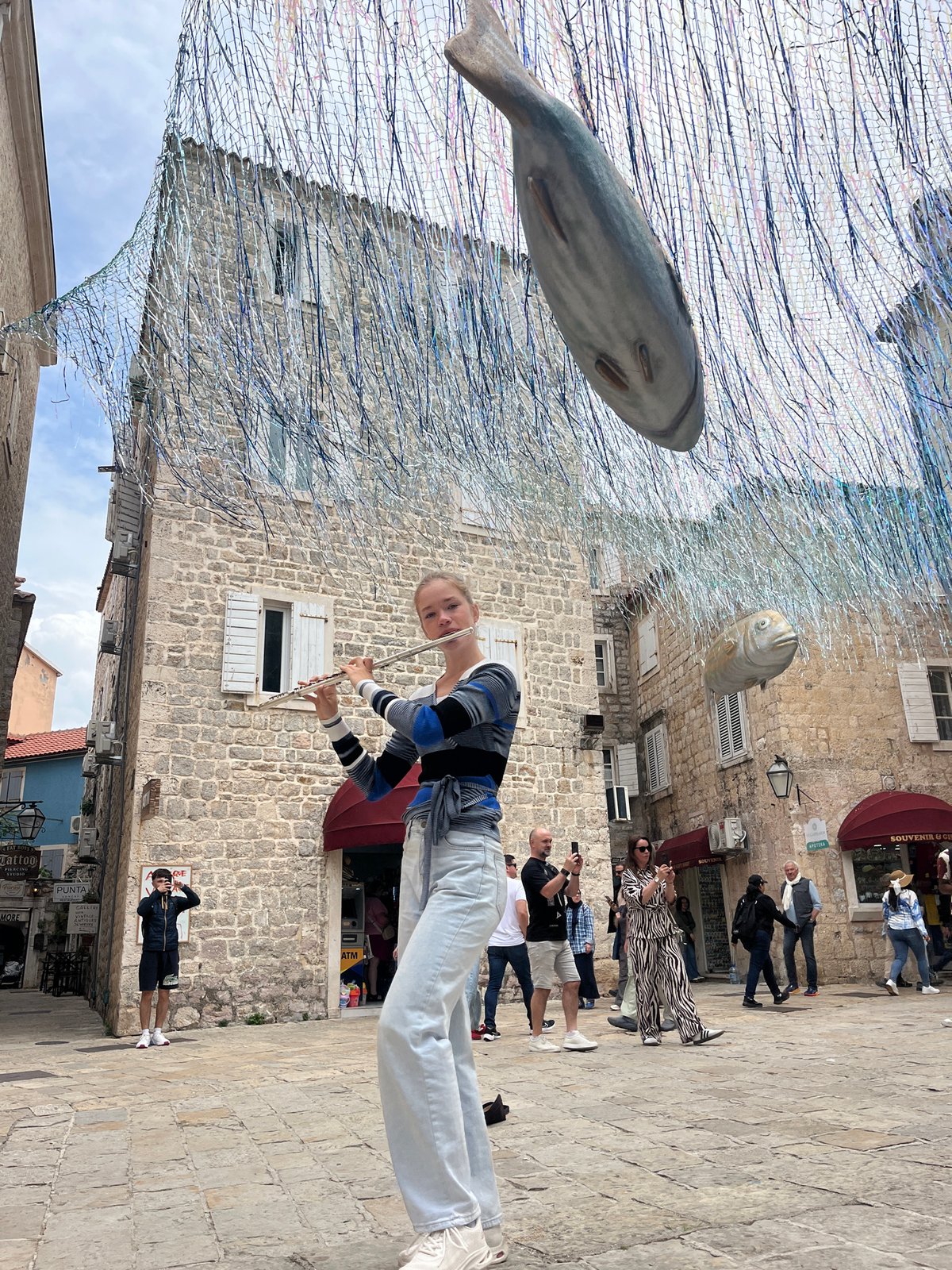 Street musician plays flute under fish sculptures in Budva