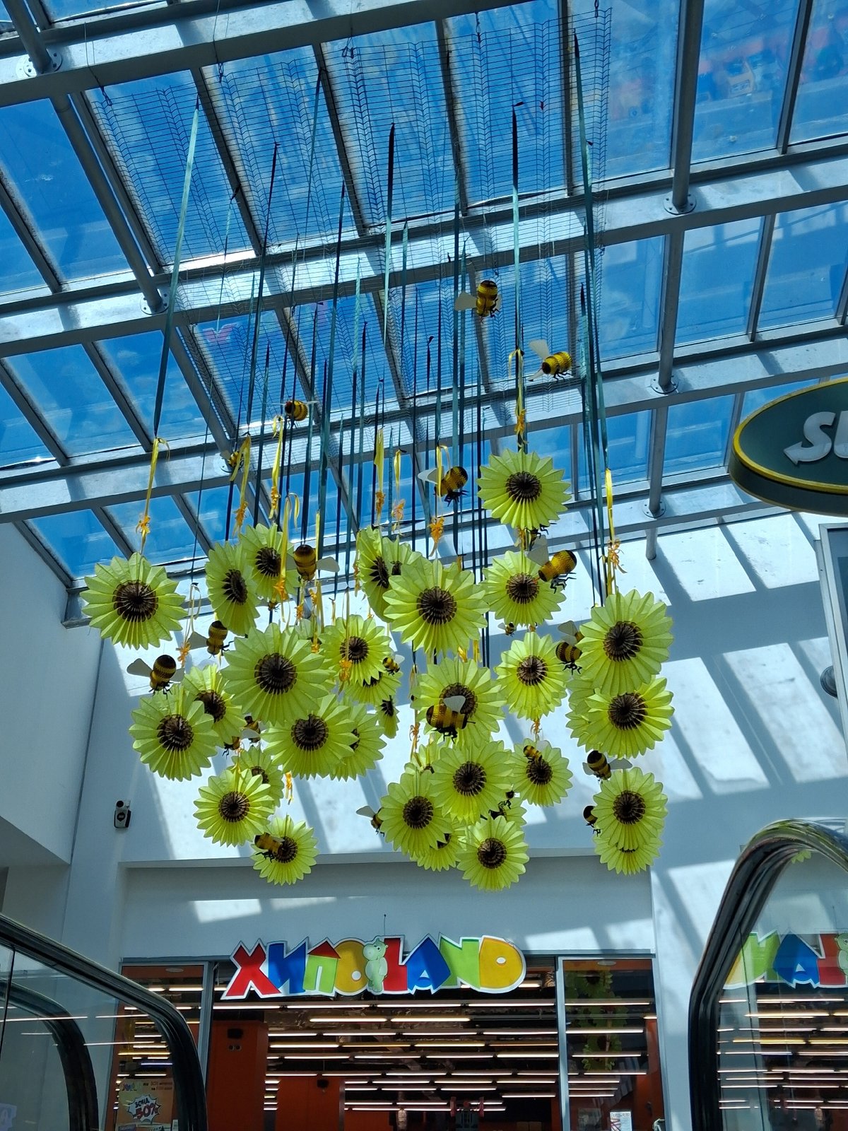 Paper sunflower decorations hang from glass ceiling in interior space