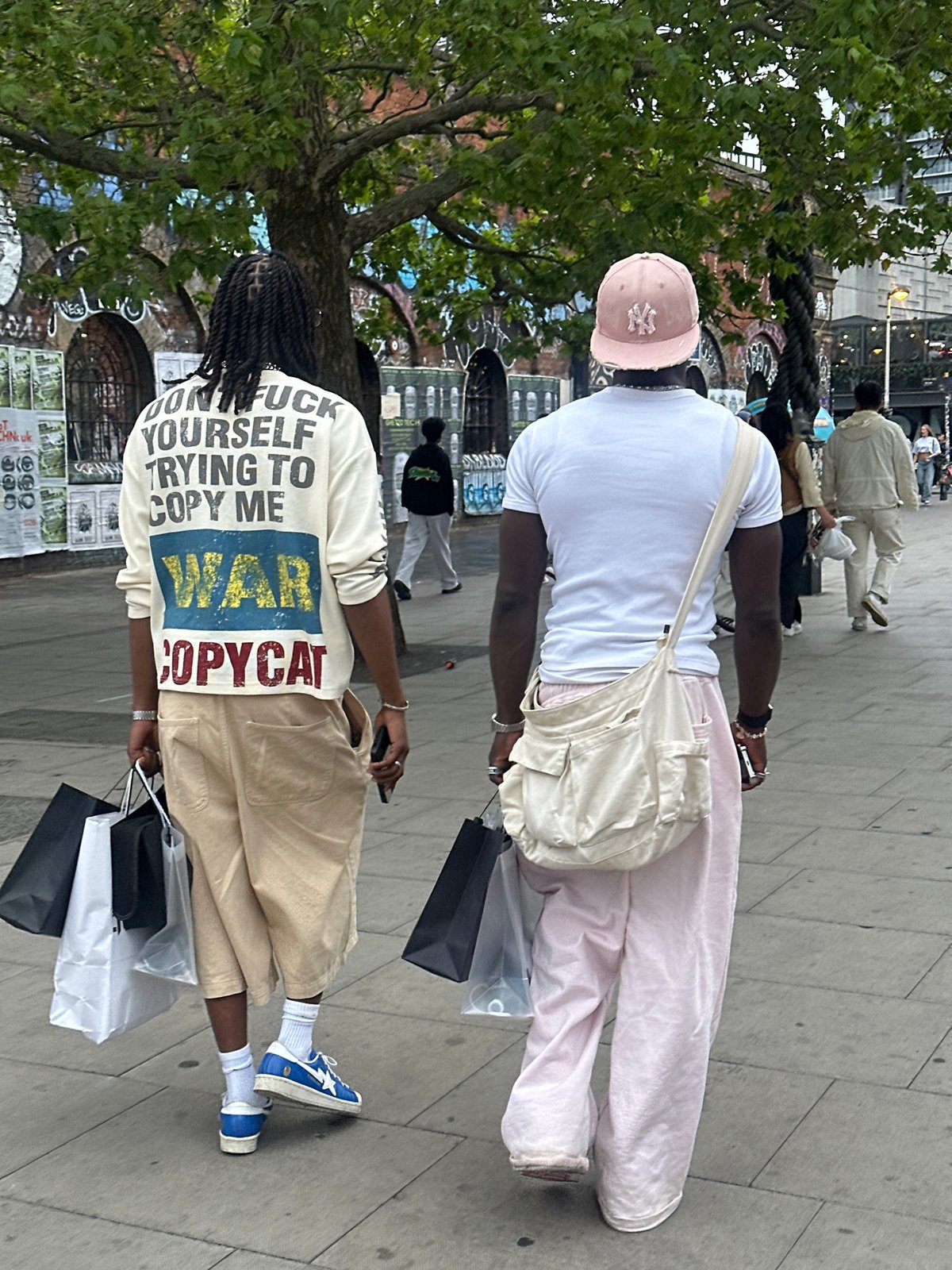 Two pedestrians walk London sidewalk, one wearing slogan shirt