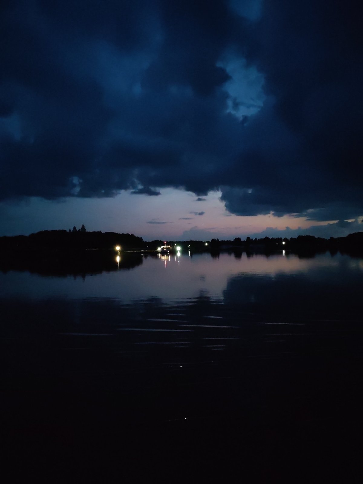 Still water mirrors distant settlement lights under a stormy Russian sky.