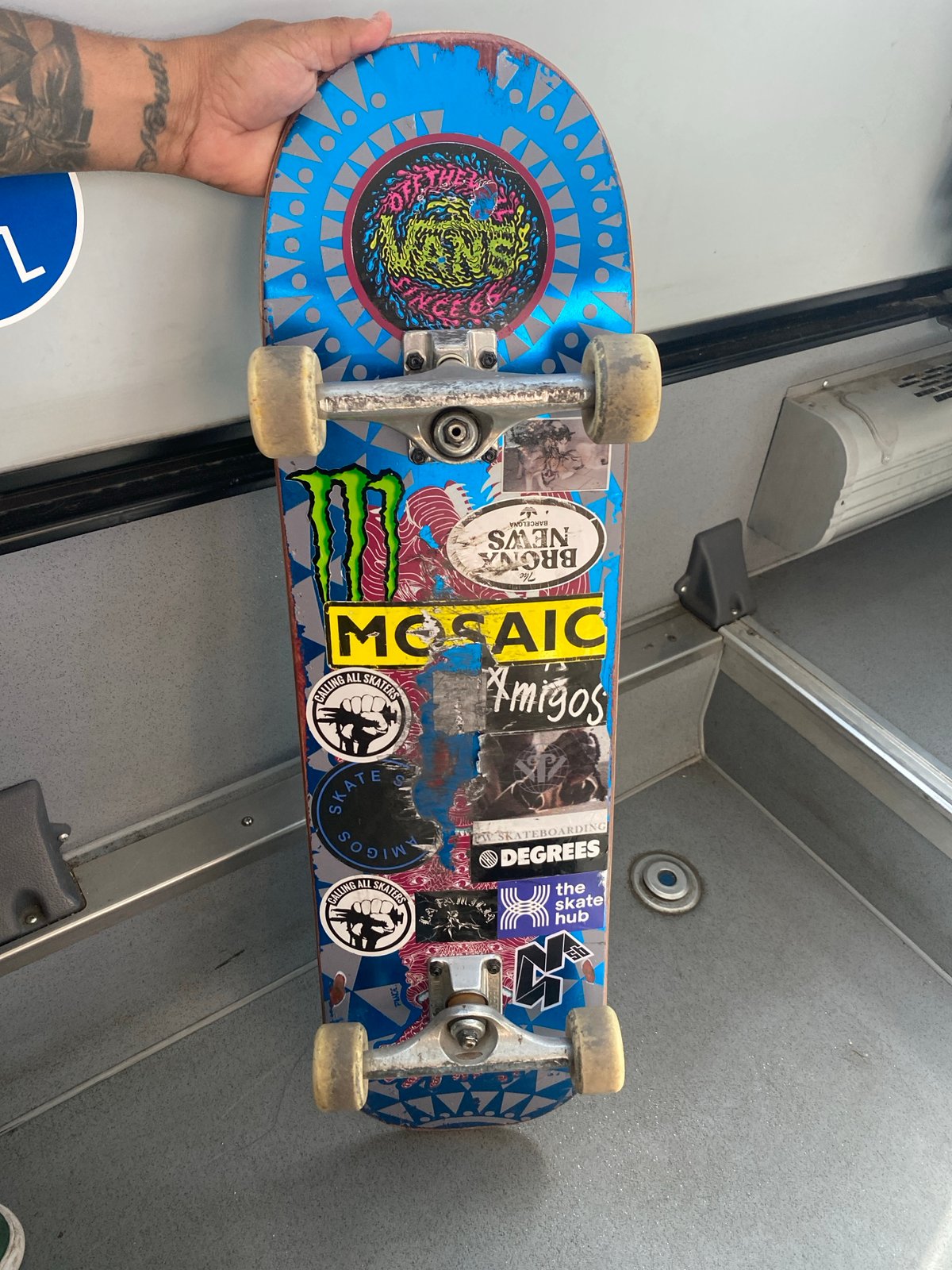 Tattooed skateboarder holds sticker-covered blue deck in Barcelona
