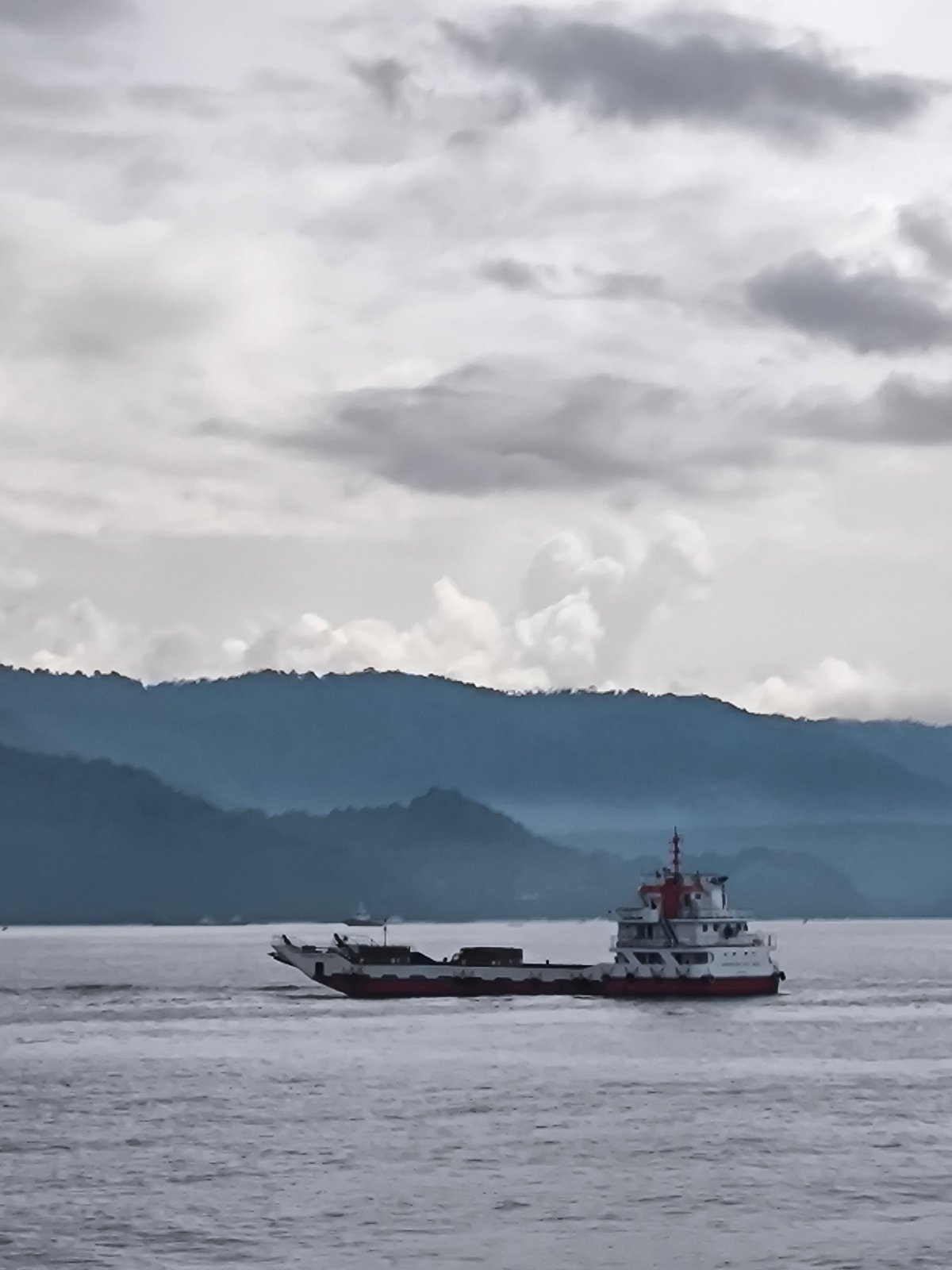 Ferry navigates Indonesian waters near mountainous coastline under overcast skies