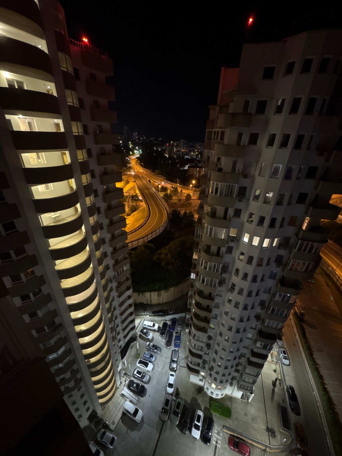 Woman at window observes Sochi nighttime cityscape from apartment
