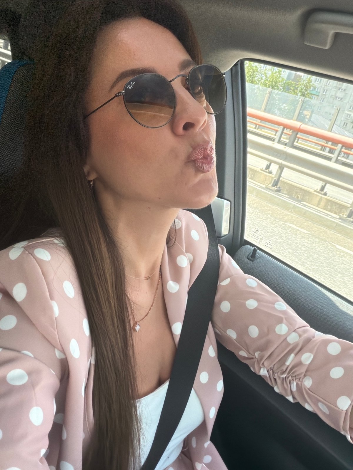 Inside a car in Riga, pink blazer, city streets rolling by.