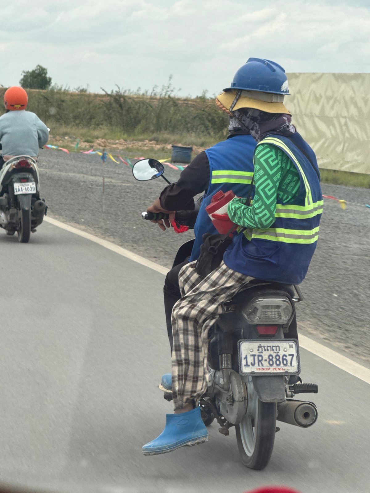 Motorcycle riders navigate Krong Ta Khmau streets, layered up against the elements