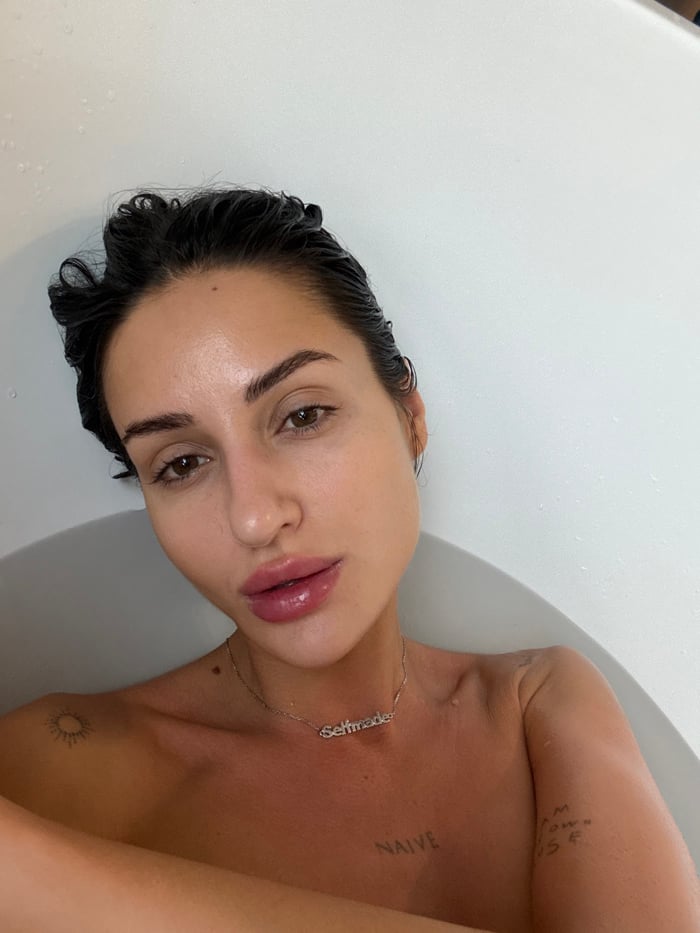 Woman observed in bathtub wearing silver necklace, tattoos visible