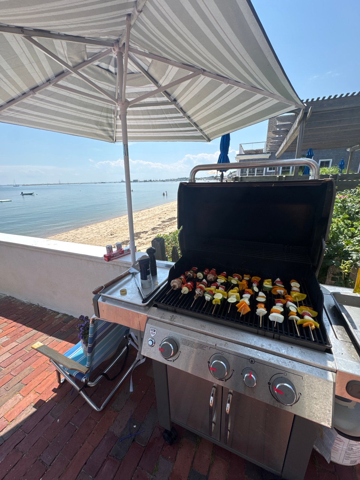 Vegetable skewers on an open barbecue grill under a striped umbrella
