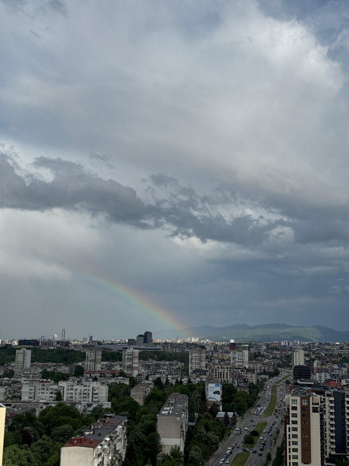 Rainbow appears over dense urban cityscape amid cloudy afternoon