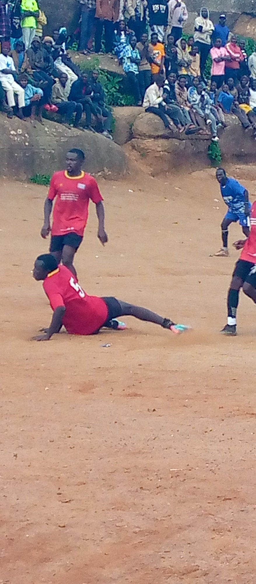 Informal soccer match on a dirt field, crowd on rocky hillside