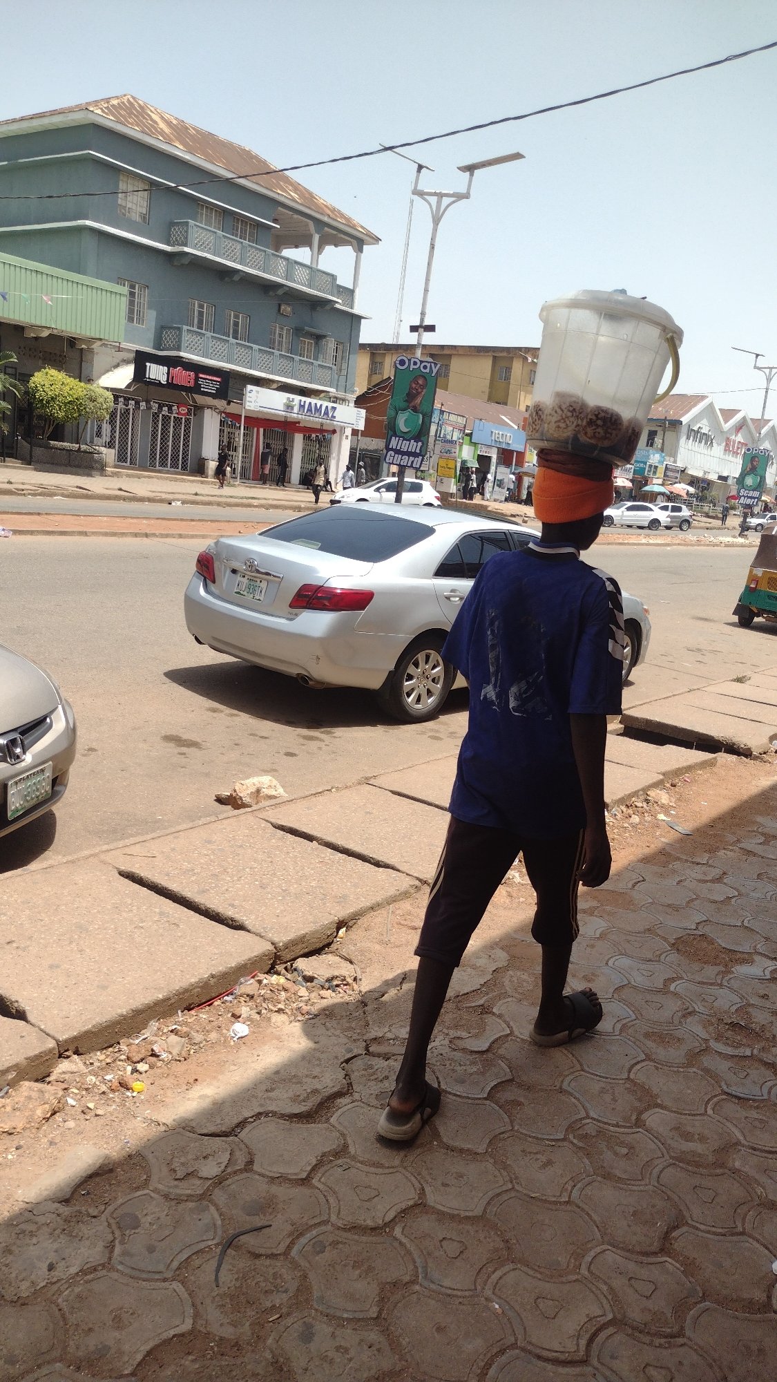 Person carries container on head along Jos sidewalk