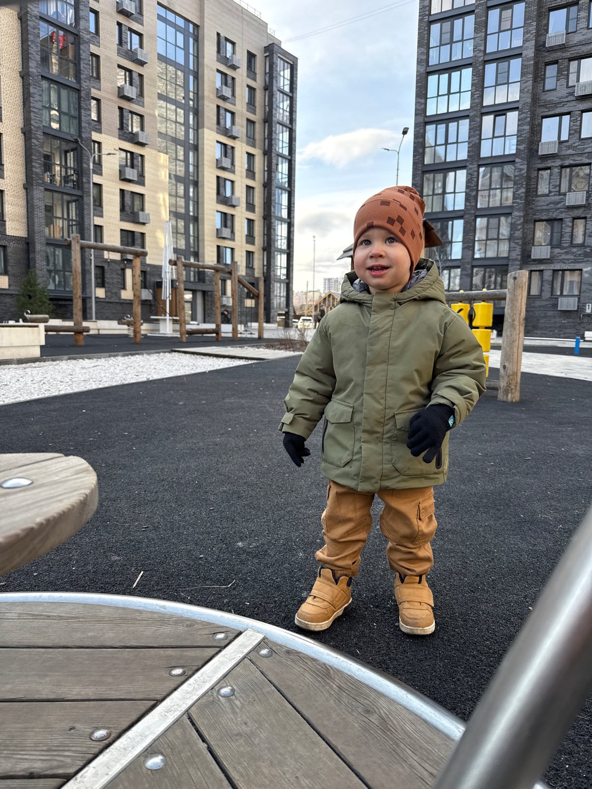 Toddler in winter clothing stands on playground surface near building