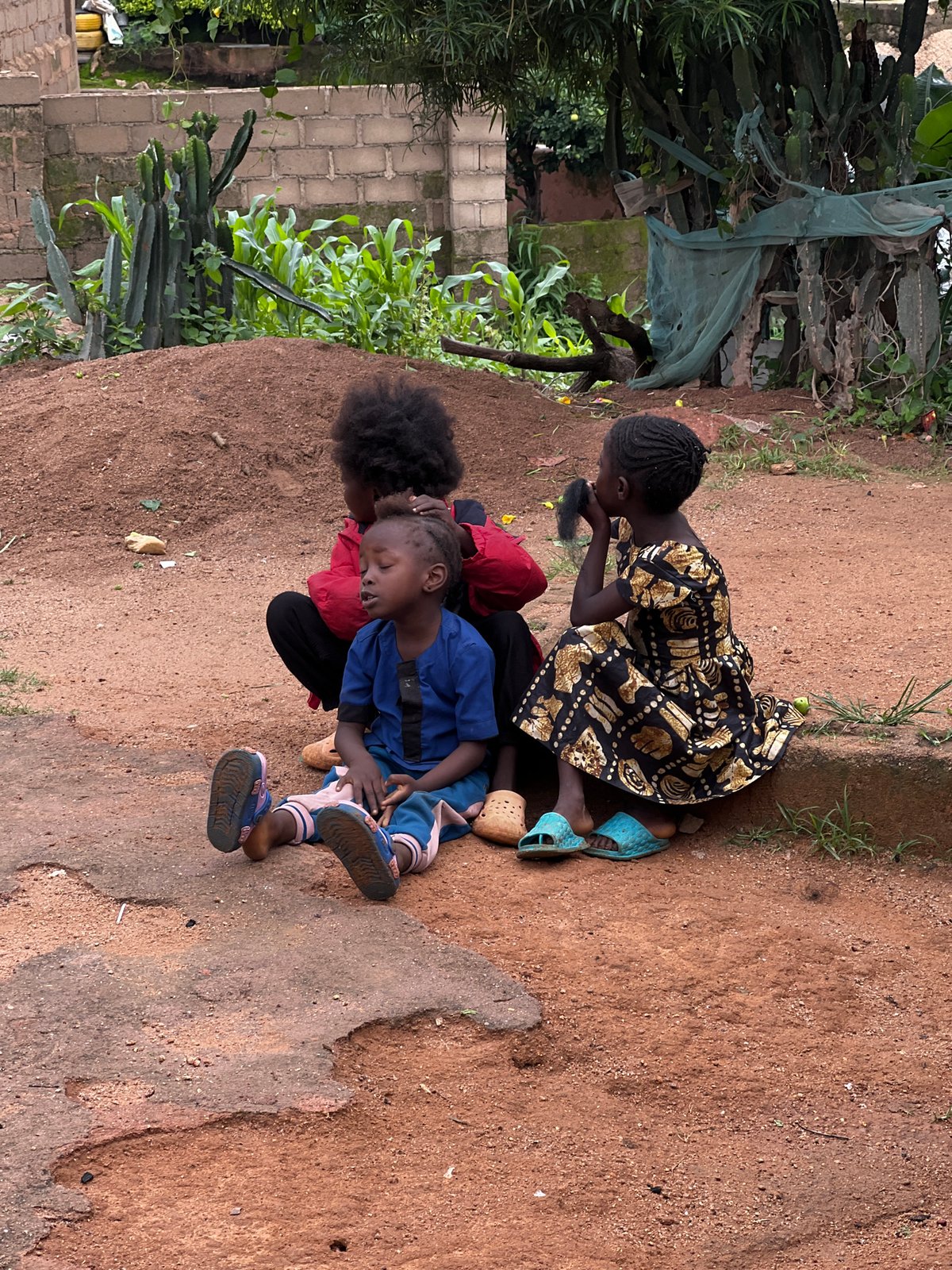 Kids hanging out on a dirt patch in Jos, one getting a hair braid