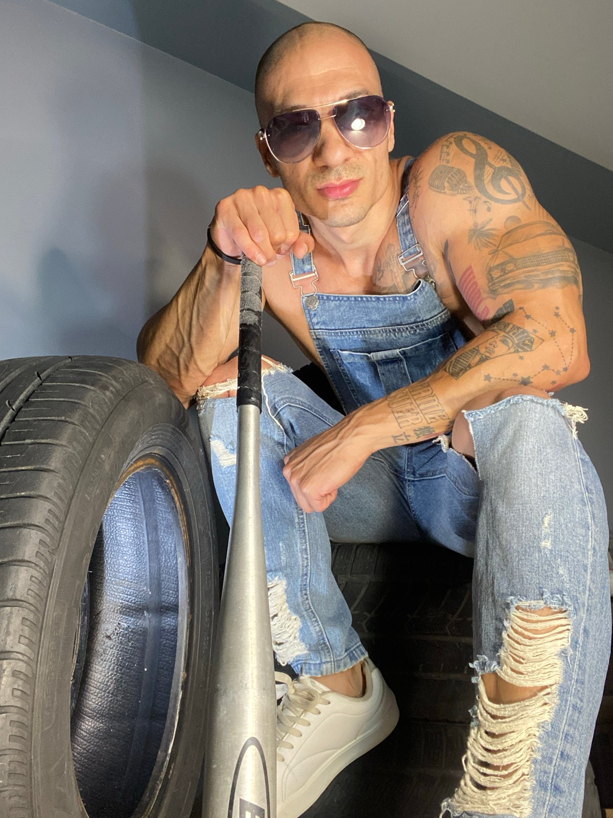 Tattooed figure poses with baseball bat among stacked tires in Pleven