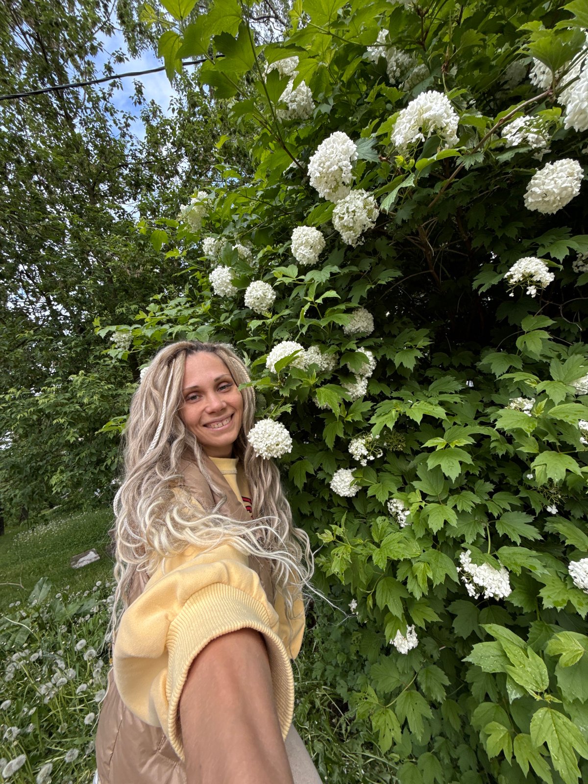 Woman in yellow sweatshirt photographed outdoors with flowering bushes