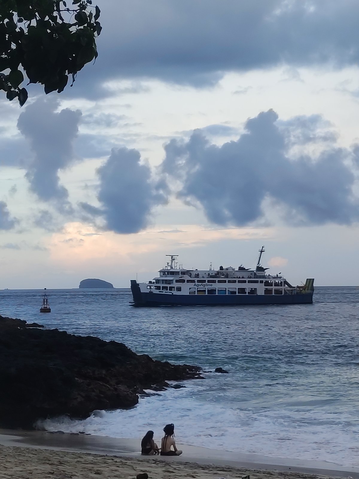 Two people sit on beach as ferry passes rocky coastline