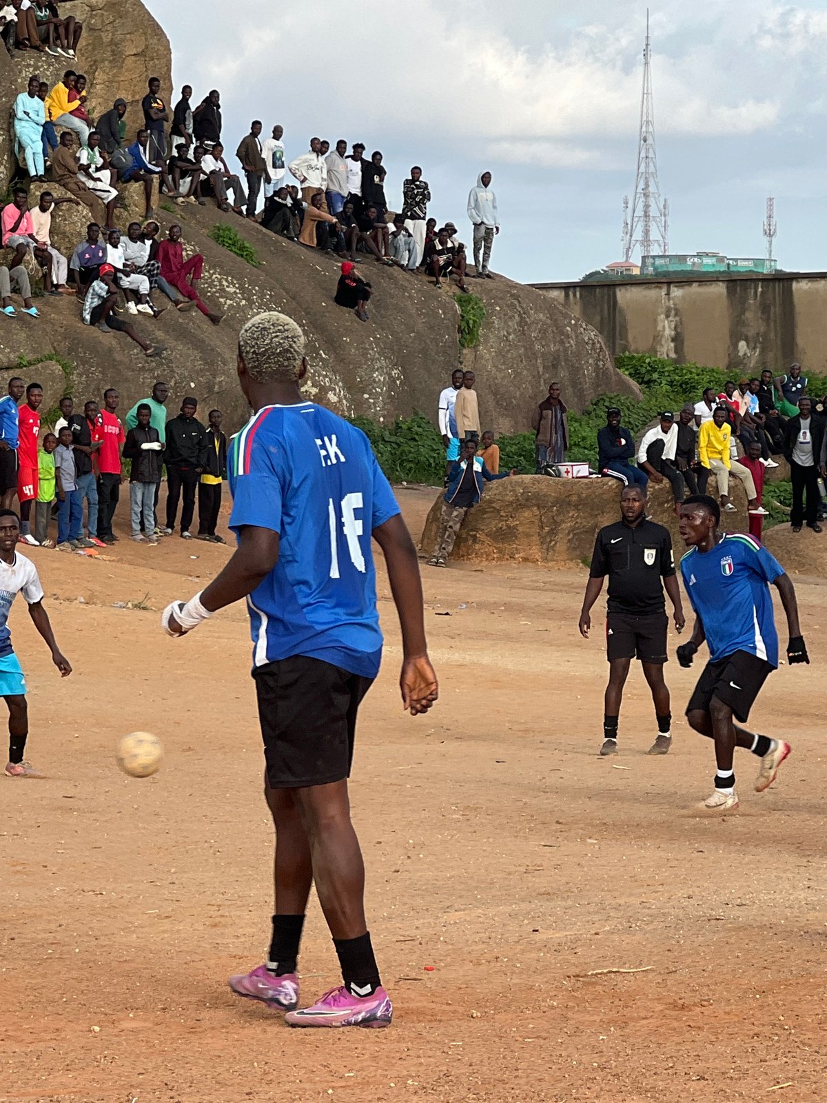 Pickup football on a rocky Jos field, crowds watching from above