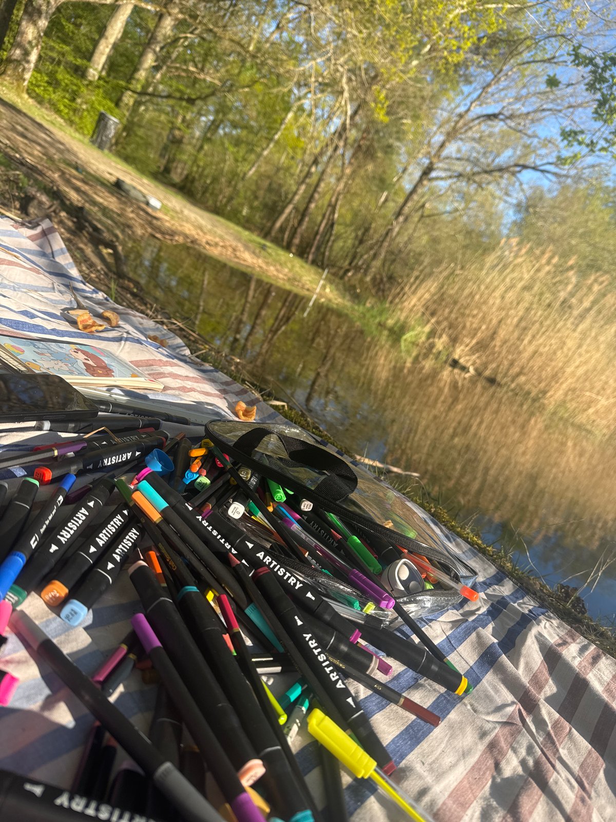 Artist with markers sits on blanket by water in Combreux