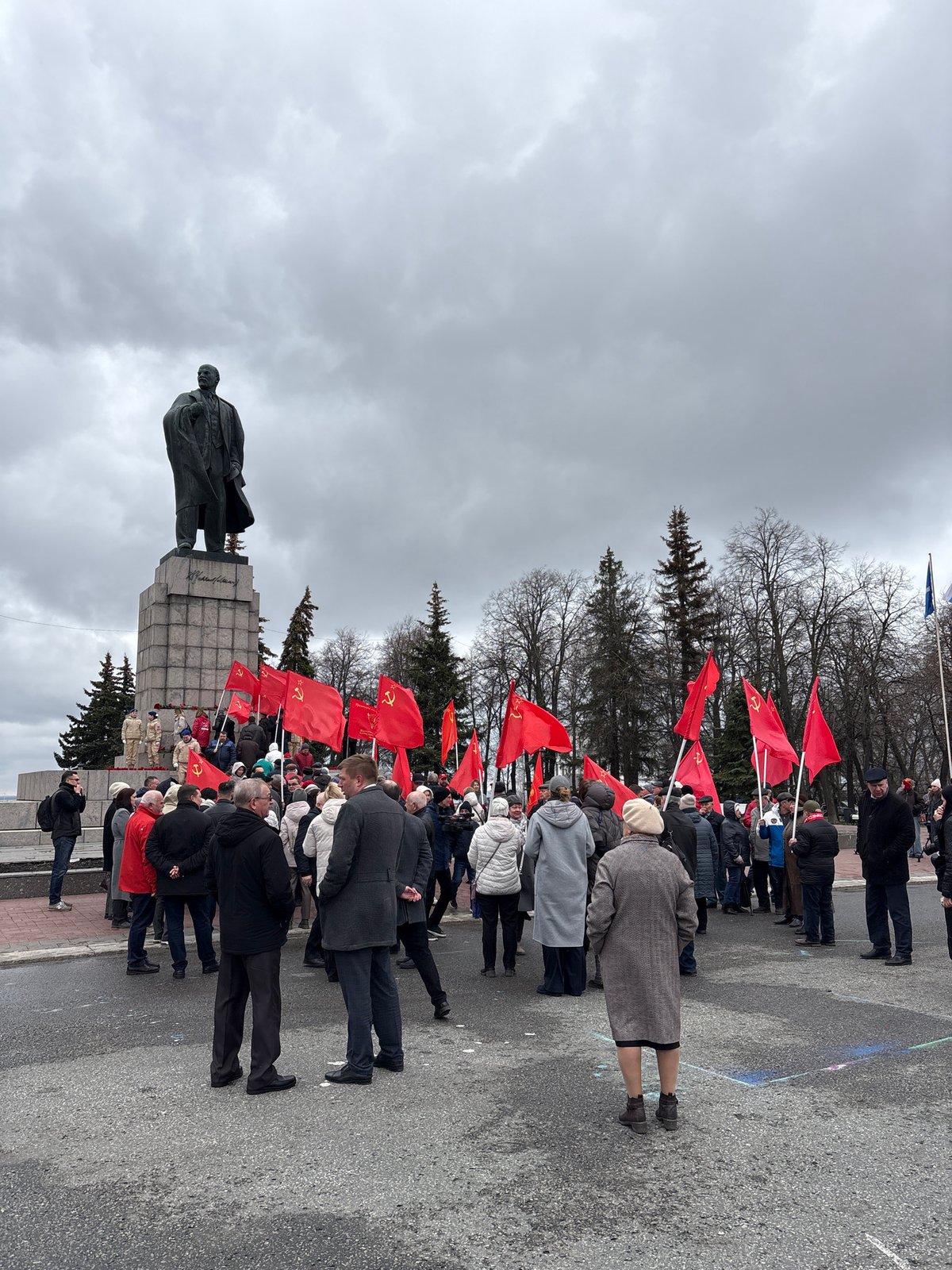 Elderly crowd gathers at Lenin statue with red flags