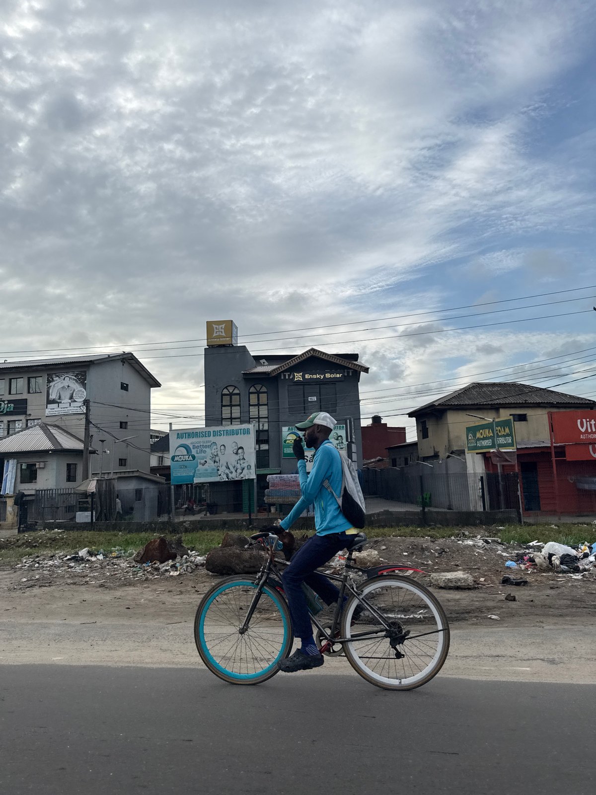Cyclist rides blue-rimmed bicycle on debris-lined road in Nigeria