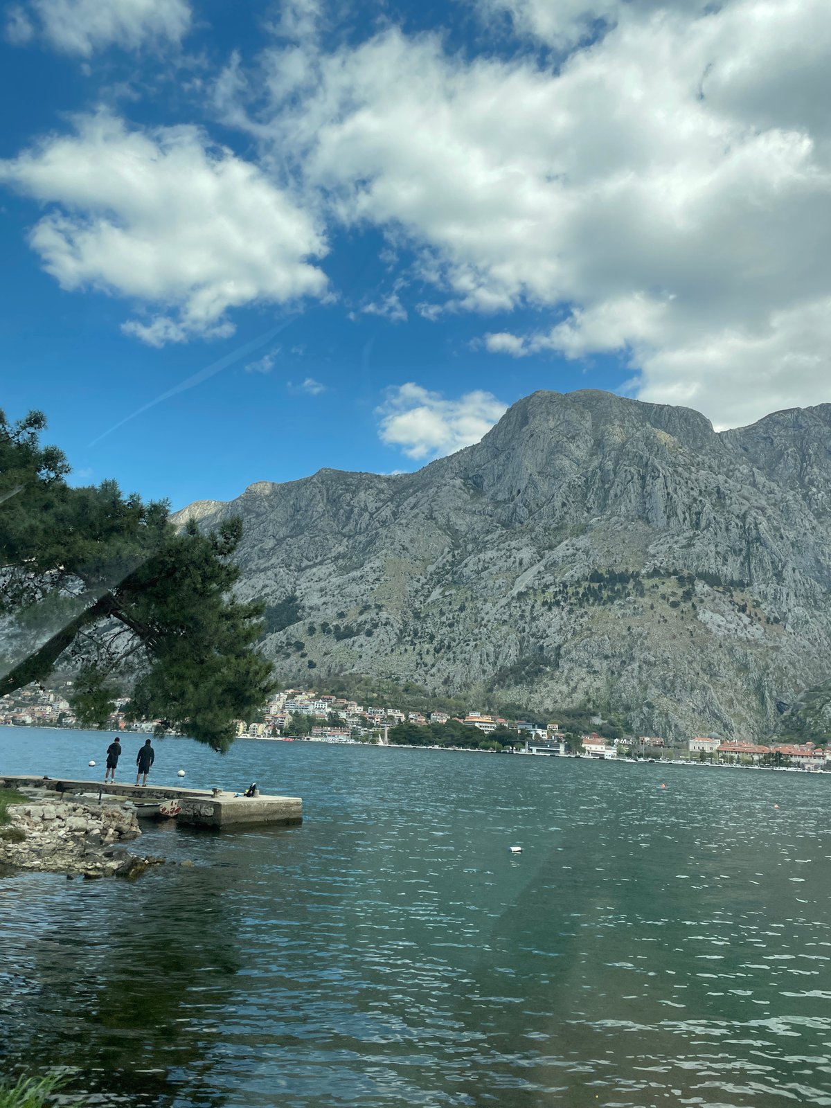 People gather at Škaljari waterfront amid mountain backdrop