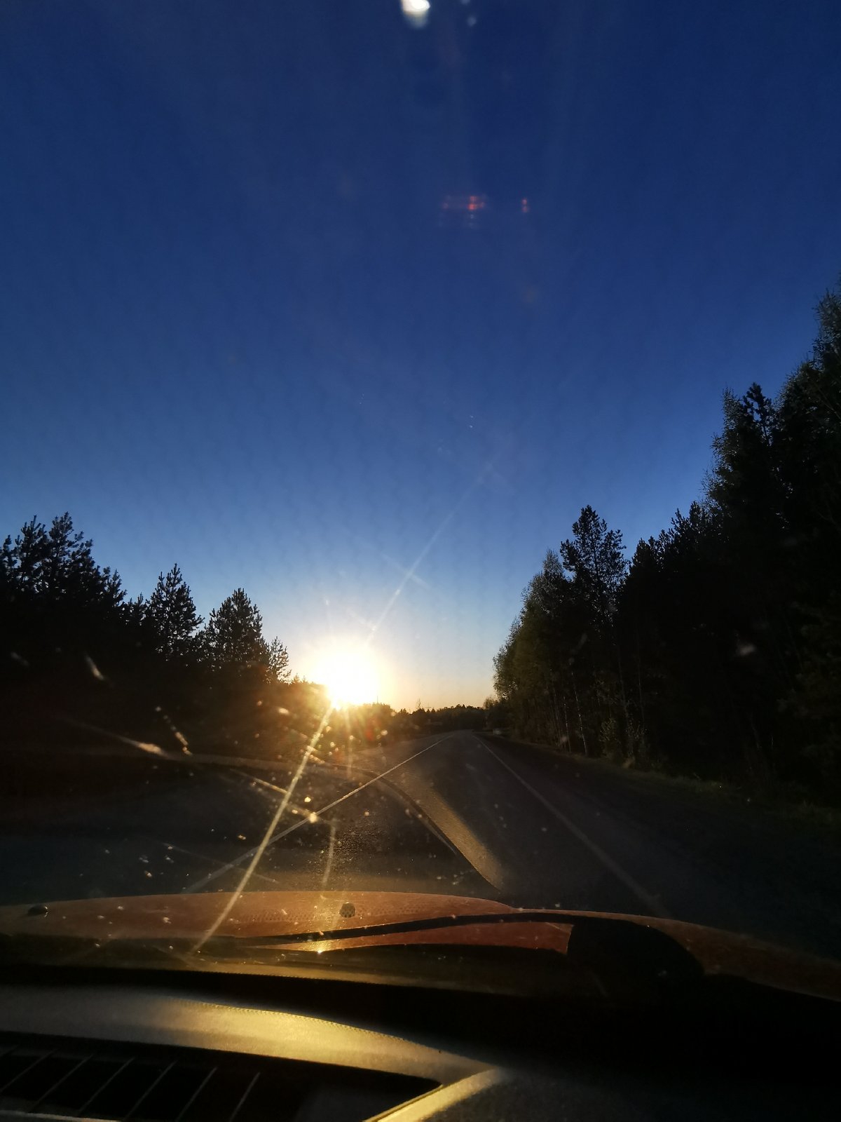 Vehicle travels tree-lined road toward colorful sky in Chaikovsky