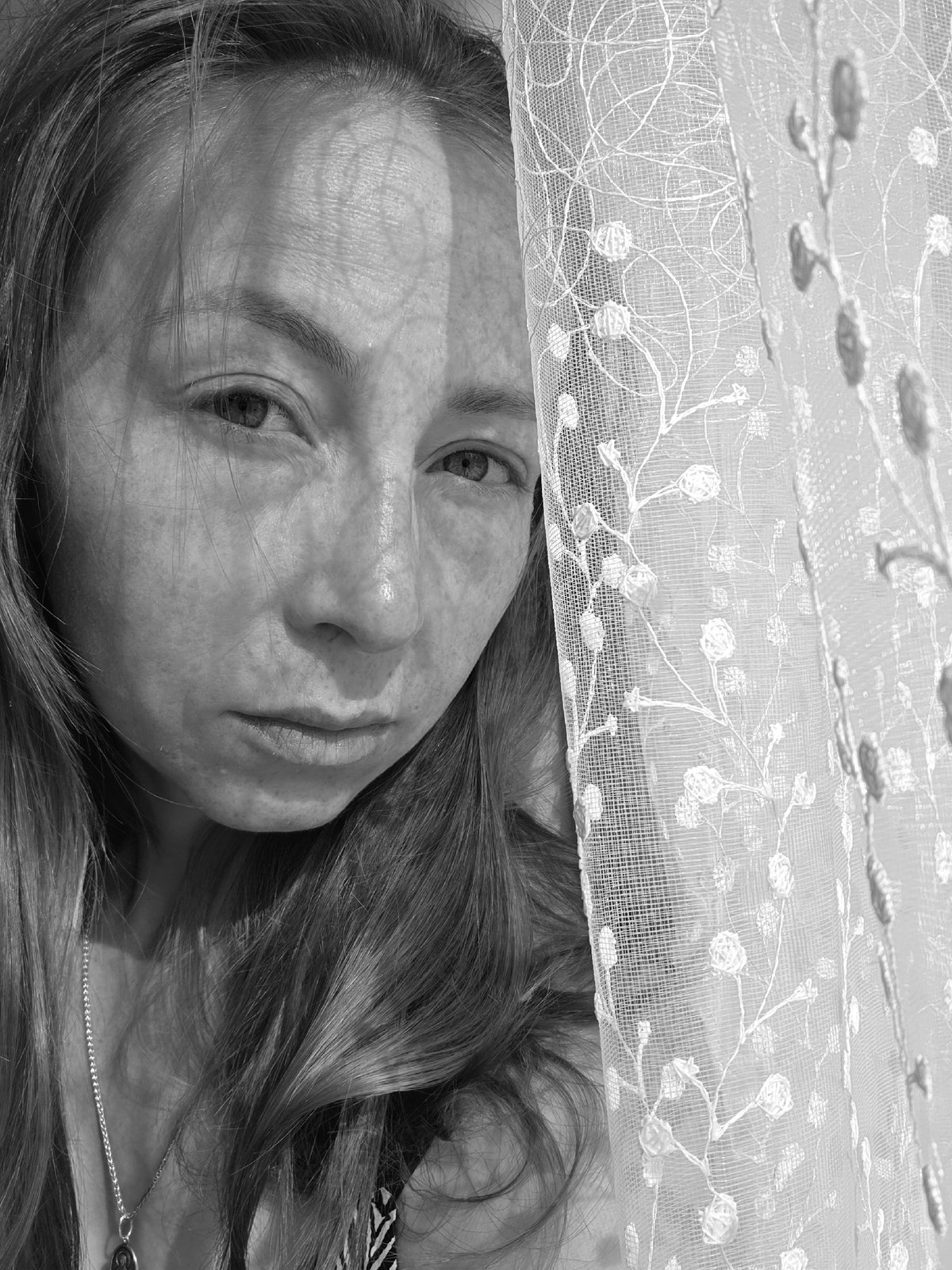 Woman photographed in intimate indoor setting near lace-curtained window