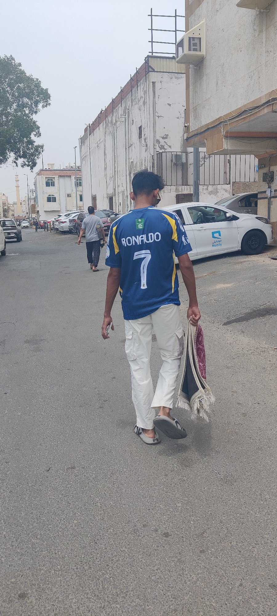 Pedestrians walk Jeddah street, man carries prayer rug