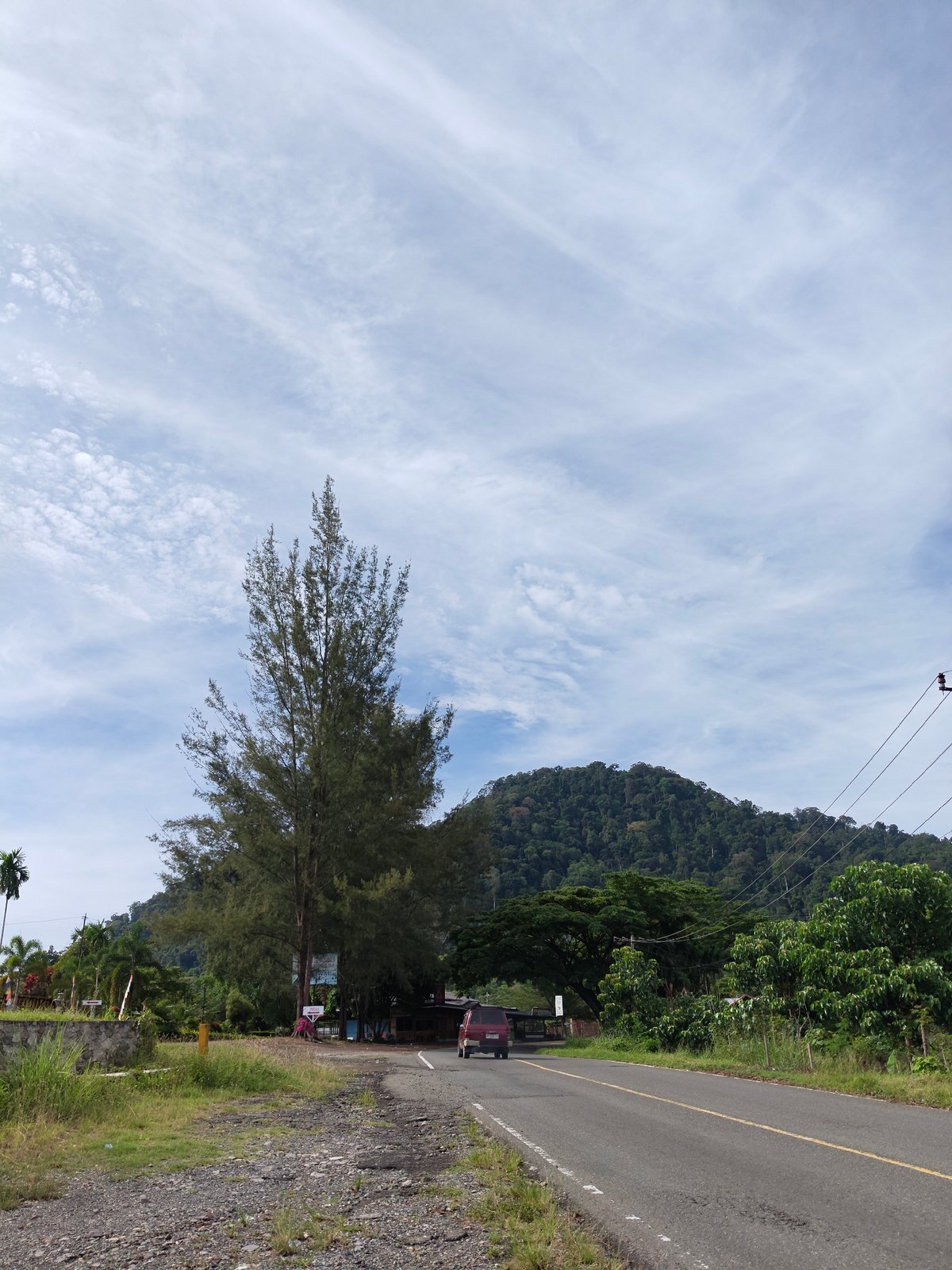 Maroon car travels Indonesian road past tree and building