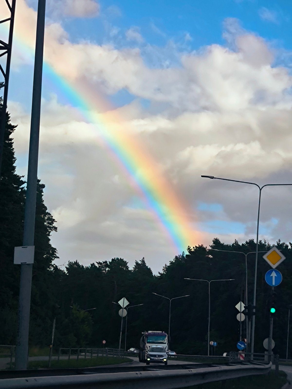 Rainbow arches over a Riga road on a partly cloudy afternoon.