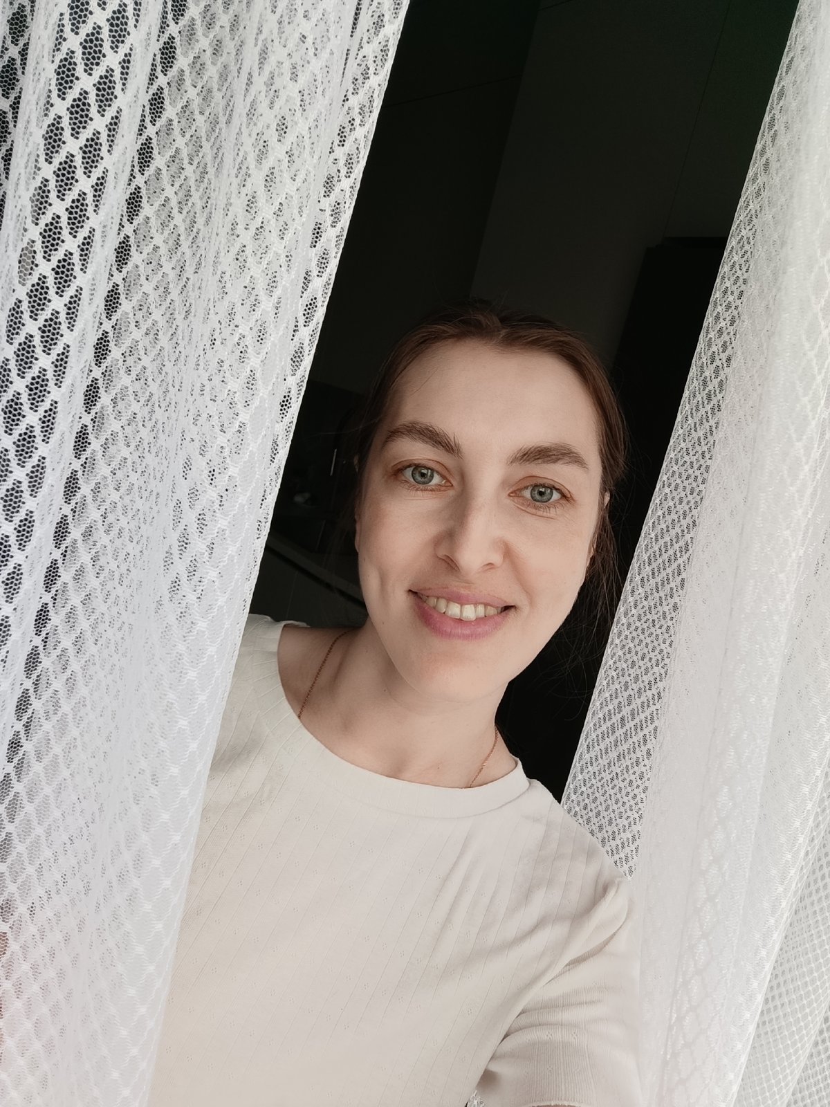 Woman photographed smiling behind lace curtains in Chaikovsky, Russia